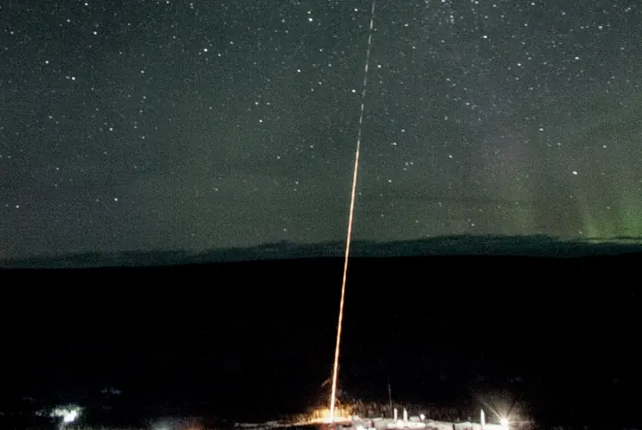 A rocket's trail rising into a field of stars above a dark horizon